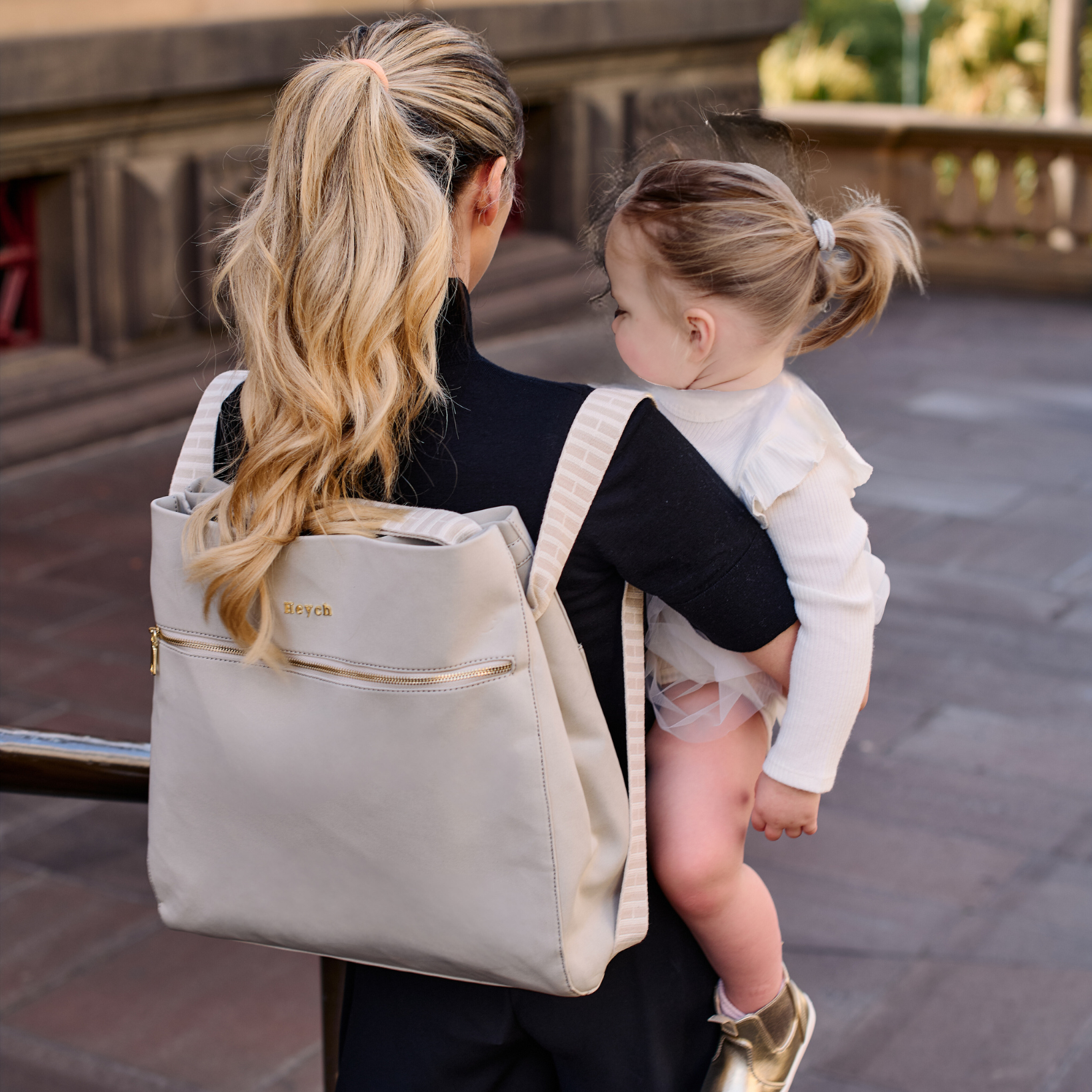 Mother carrying toddler with stone neoprene baby bag worn as a backpack