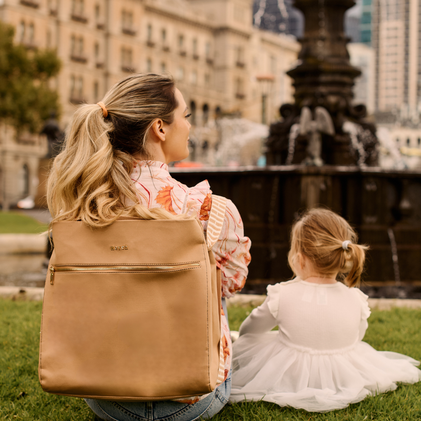 Mother wearing tan neoprene bag as a backpack sitting next to her toddler outside